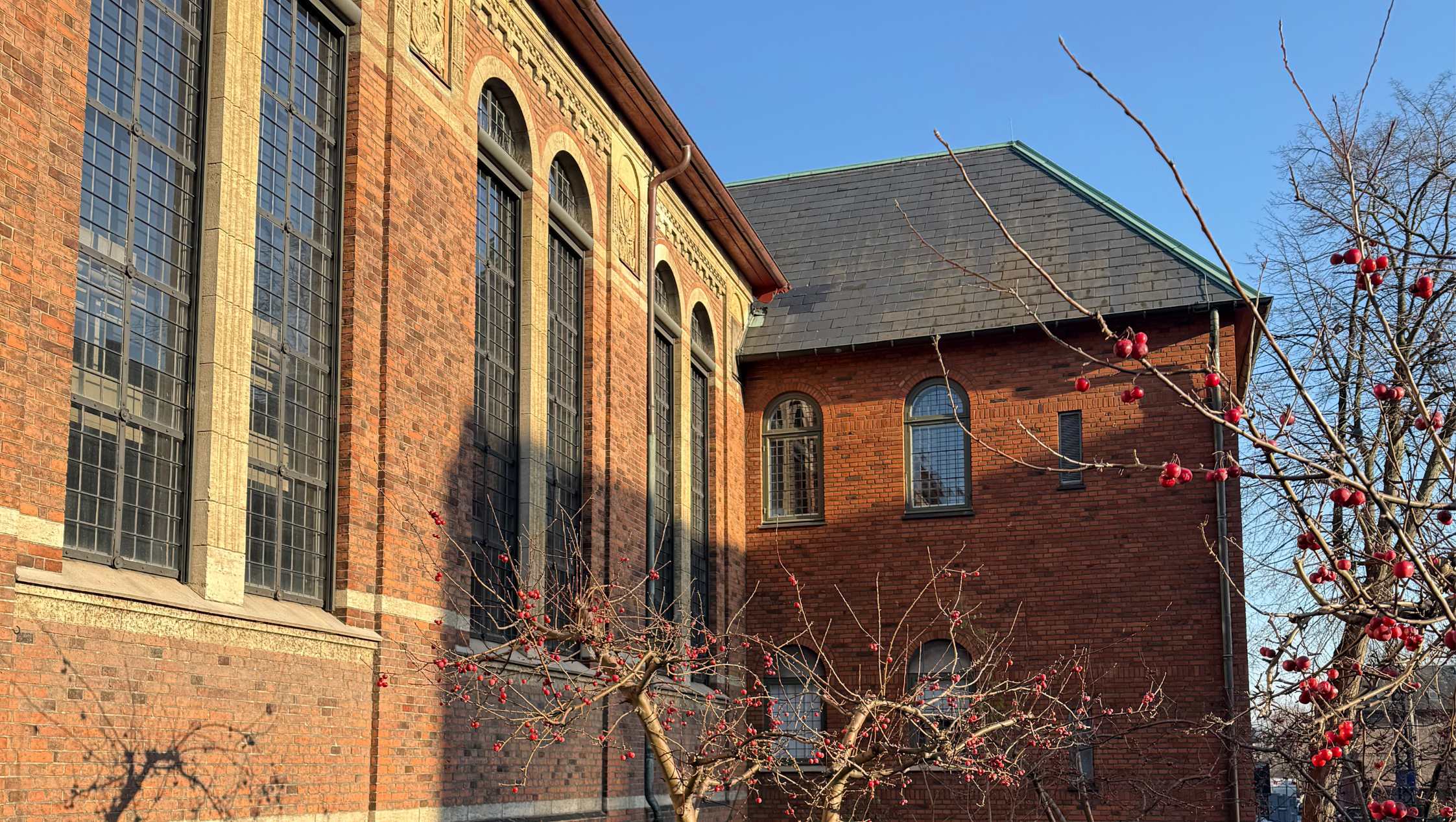 Sions Kirkes facade med solskin og blå himmel i baggrunden og røde knopper på træerne i forgrunden